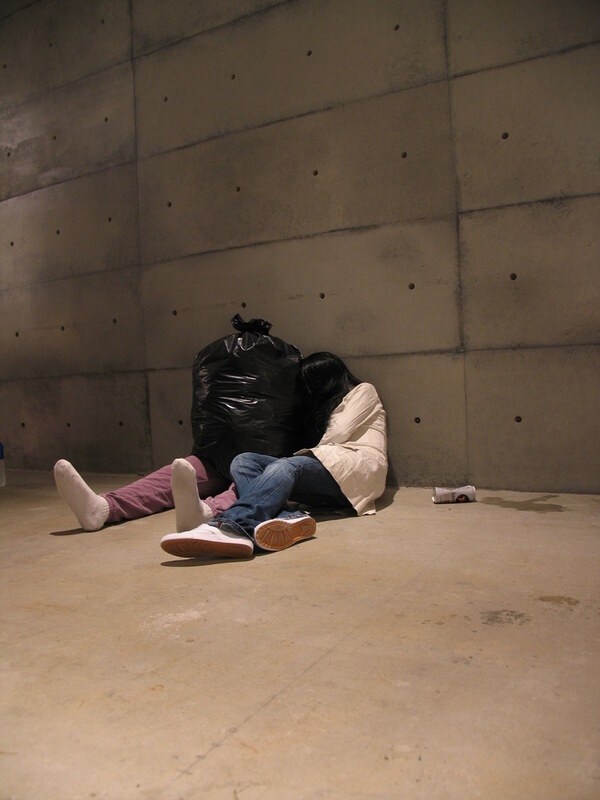 Figure slumped against a wall with a garbage-bag figure